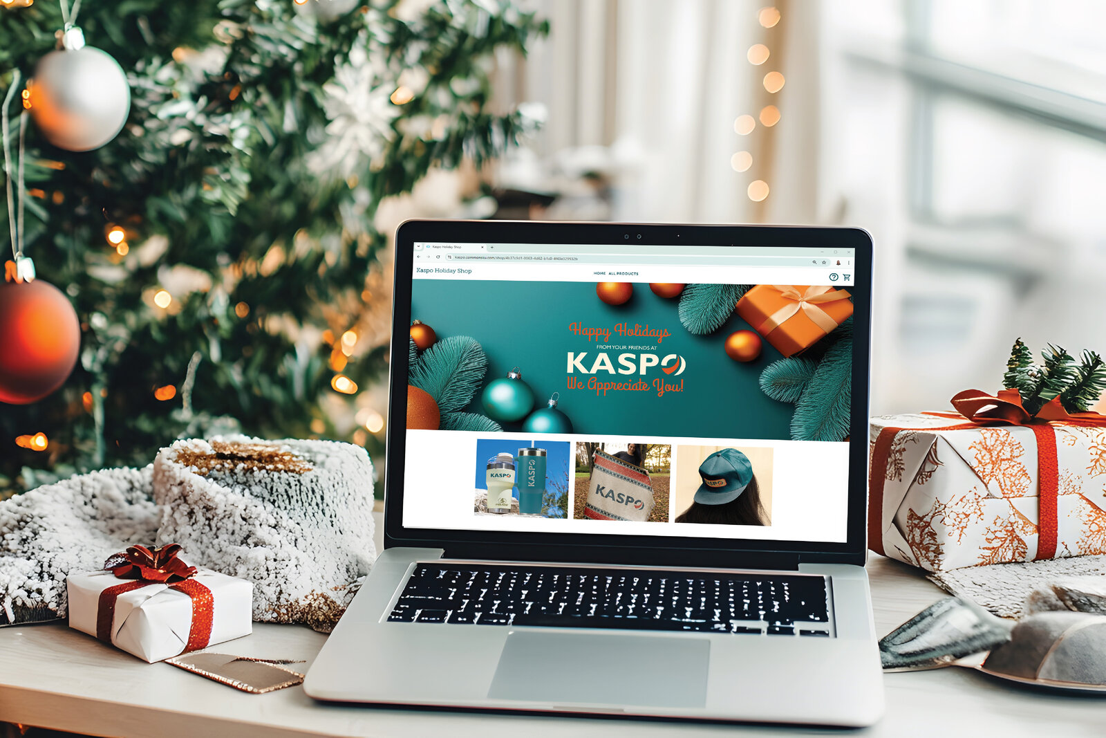 A laptop on a holiday-decorated desk showing the Kaspo Holiday Shop online gift store, with branded tumblers, a blanket, and a cap.