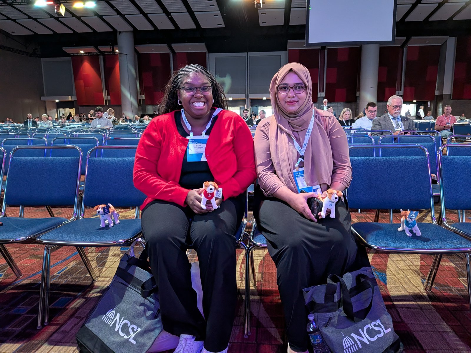 Two smiling attendees seated in the ballroom, each holding a plush Nora dog, with NCSL tote bags at their feet.