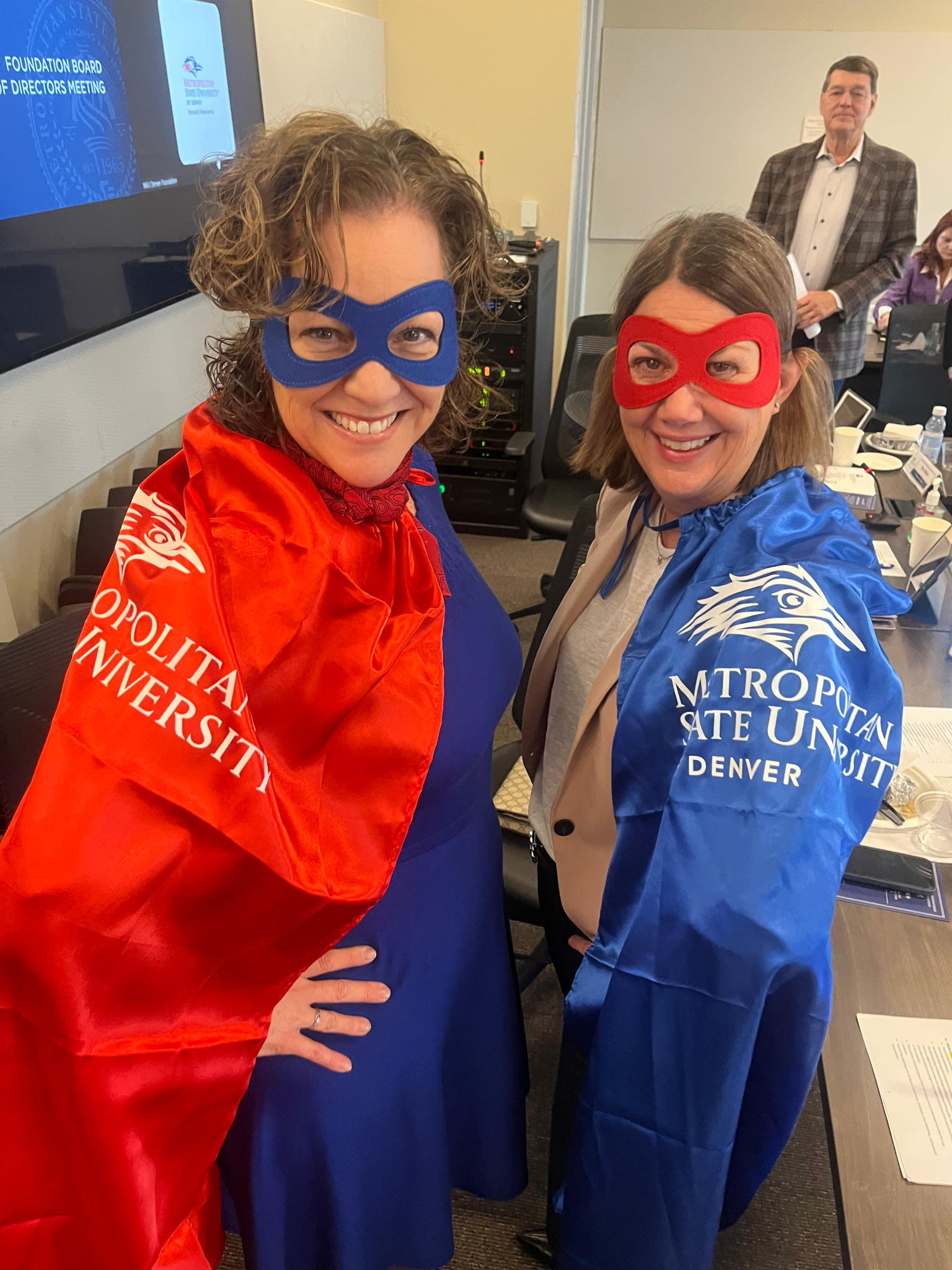 Two board members wearing custom branded satin capes and felt masks at a university foundation meeting.
