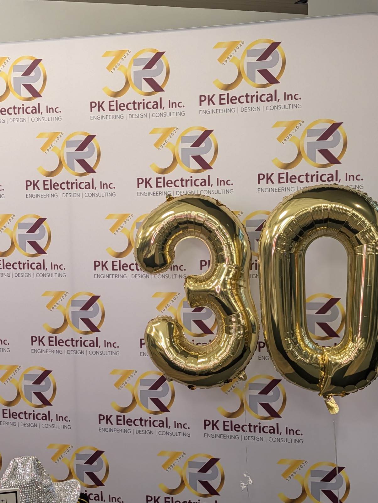 A step-and-repeat backdrop of PK Electrical 30-year logos with giant gold foil balloons spelling 30.