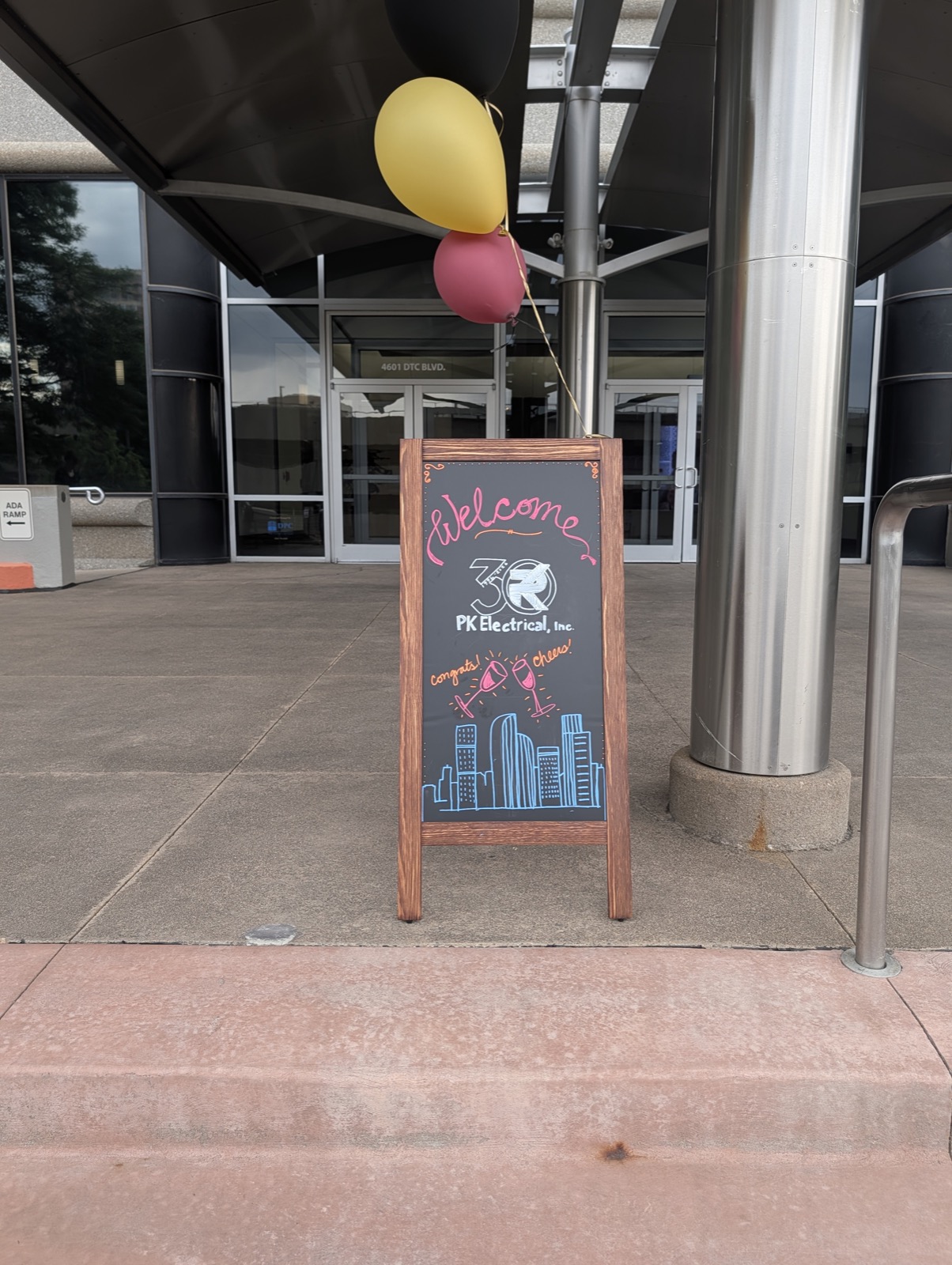 A hand-lettered chalkboard sign reading Welcome, 30, PK Electrical Inc, with a drawn Denver skyline and balloons at the building entrance.