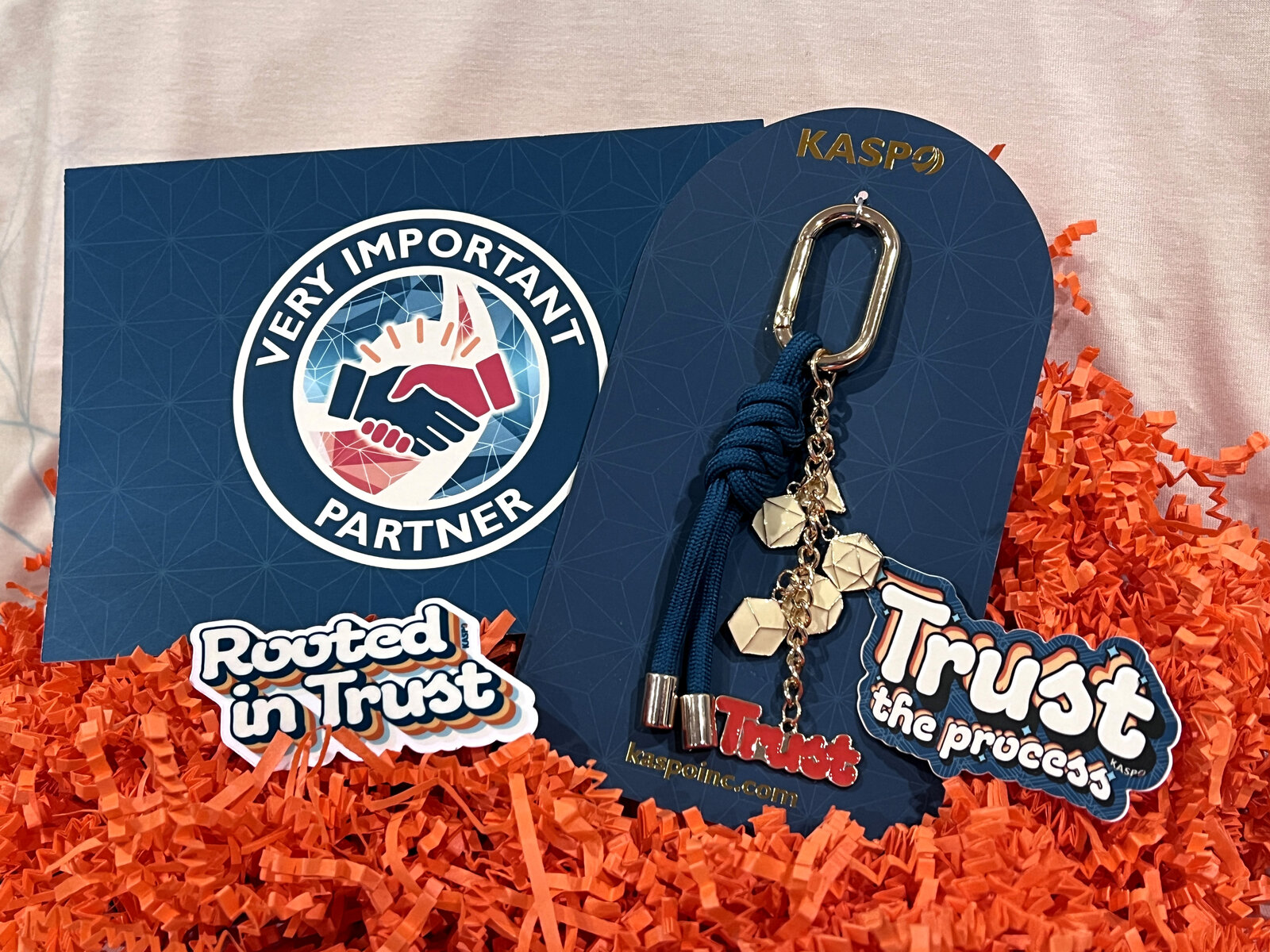 A Kaspo VIP partner mailer on orange crinkle paper: a Very Important Partner card, a trust charm keychain on a branded card, and stickers reading Rooted in Trust and Trust the process.